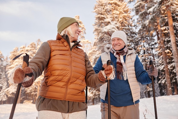 Low Angle Portrait eines glücklichen Seniorenpaares, das mit Stöcken im Winterwald spazieren geht und lächelt