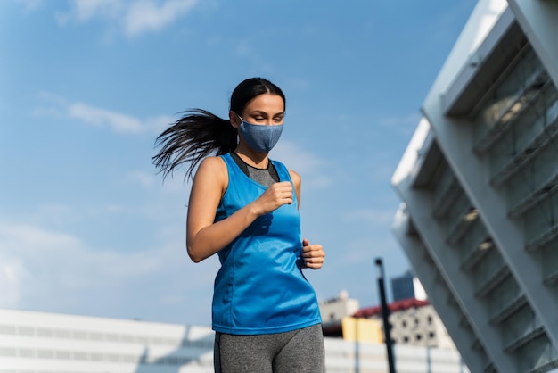 Low Angle View der jungen glücklichen Sportlerin mit Schutzmaske beim Aufwärmen des Lauftrainings auf der Straße Fröhliches Mädchen, das ihr Sportaktivitätskonzept genießt