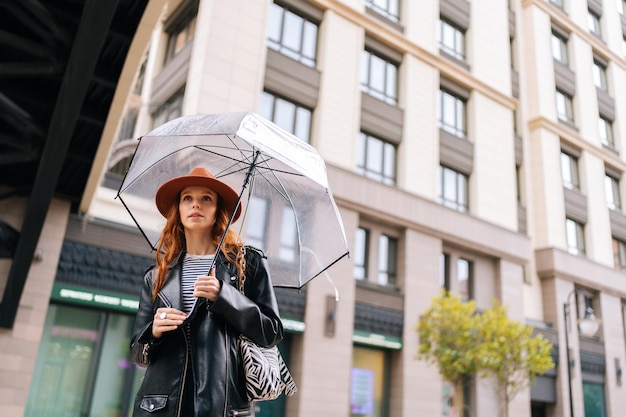 Lowangle-Ansicht einer hübschen rothaarigen jungen Frau mit modischem Hut, die bei regnerischem Regen mit transparentem Regenschirm auf der Stadtstraße wartet und wegschaut