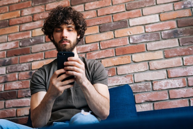 Lowangle-Ansicht eines ernsthaften bärtigen jungen Mannes, der auf dem Smartphone-Bildschirm beim Surfen im drahtlosen Internet auf dem Gerät sitzt, das zu Hause auf dem Sofa sitzt