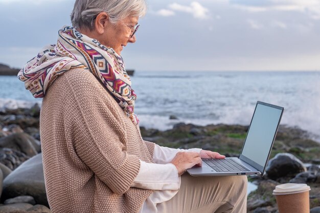 Ältere aktive Frau, die mit einem Laptop am Strand sitzt und einen Schal trägt Ältere attraktive Frau in Fernarbeit, die den Sonnenuntergang am Meereshorizont über Wasser genießt