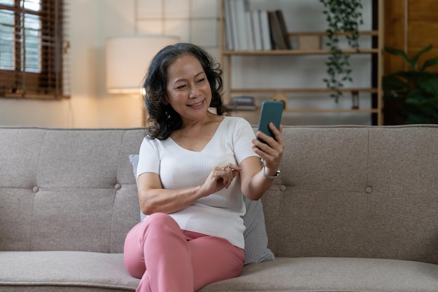 Ältere Asiatin, die Handy benutzt und zu Hause auf Sofa sitzt