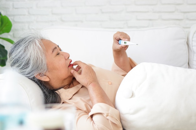 Ältere Asiatin liegt krank auf dem Sofa im Haus Nehmen Sie Medikamente gegen Fieber ein, wie vom Arzt verschrieben. und verwenden Sie ein digitales Thermometer, um die Temperatur zu überprüfen. gönnen Sie sich zu Hause