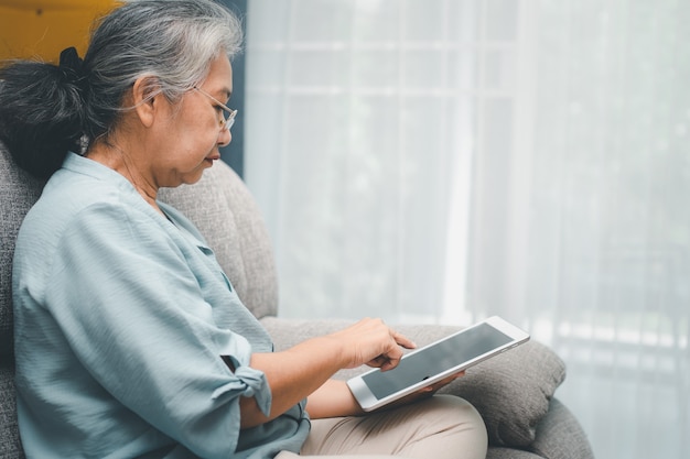 Ältere asiatische Frau, die eine Brille trägt und sich zu Hause auf einem Sofa entspannt und ein Tablet zum Lesen von Nachrichten verwendet