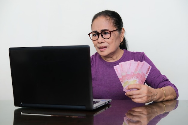 Ältere asiatische Frau, die mit verwirrtem Ausdruck auf ihren Laptop schaut, während ihre Hand Geld hält