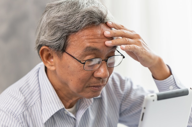 Ältere asiatische Gläser des alten Mannes Kopfschmerzen vom Verwenden und Schauen des Tablettenschirmes