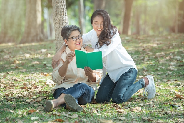 Ältere asiatische Mutter und Tochter lesen ein Buch im Wald auf grünem Rasen, das Konzept eines glücklichen Familienlebens und Familienbeziehungen, Ruhestand Leben haben Urlaub Lebensstil.