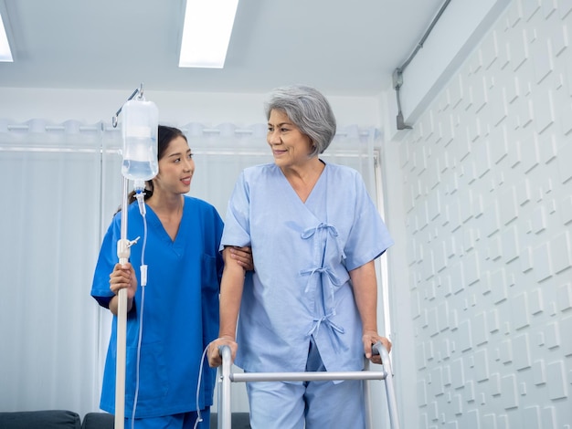 Ältere asiatische Patientin, die versucht, auf einem Laufrahmen zu gehen, wird von der Pflegekraft gehalten und sorgfältig in den Armen gestützt