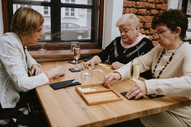 Ältere dunkelhaarige und hellhaarige Frauen mit Make-up in schwarzen und weißen Jacken, die im Café Domino spielen, mit Freunden, die ein Treffen am Tisch mit Gläsern Wein oder Champagner darauf feiern