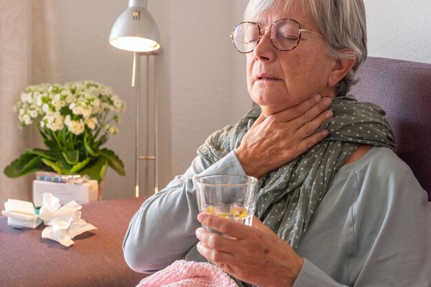 Ältere erwachsene kaukasische Frau mit Halsschmerzen und Fieber als saisonale Grippe oder Pollenallergie auf dem Sofa zu Hause, während sie ein Glas Wasser trinkt