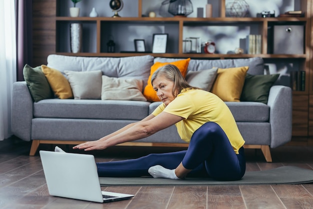 Ältere Frau beim Fitnesstraining auf dem Boden zu Hause sitzend mit Laptop für Online-Sportgruppenaktivitäten