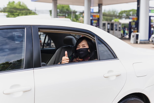 Ältere Frau des Coronavirus-Pandemiekonzepts mit dem Schutzmasken-Daumen oben sitzen in einer Auto-Straßenfahrtreise.