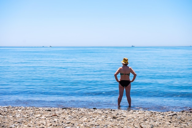 Ältere Frau, die an einem sonnigen Sommertag allein vor dem Mittelmeer an einem Strand steht