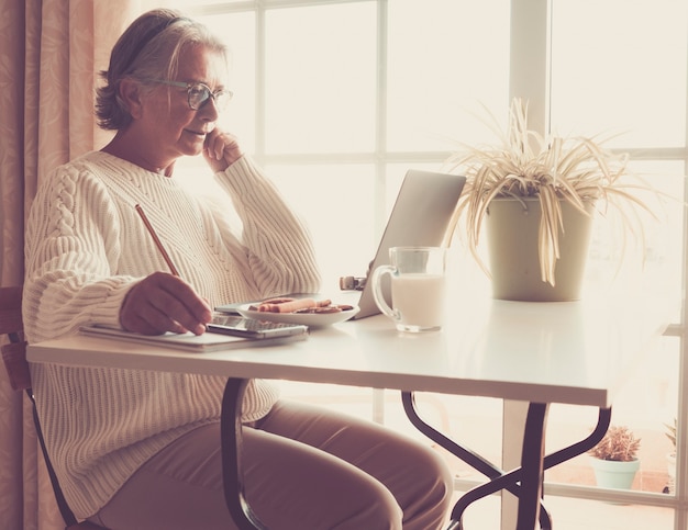 Ältere Frau, die an Laptop arbeitet. Neben ihr ein Glas Milch. Weißer Schreibtisch. Helles Licht aus dem Fenster. Alte Leute mit neuer Technik