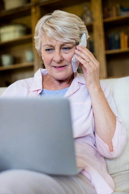 Ältere Frau, die auf Mobiltelefon spricht, während Laptop im Wohnzimmer verwendet wird