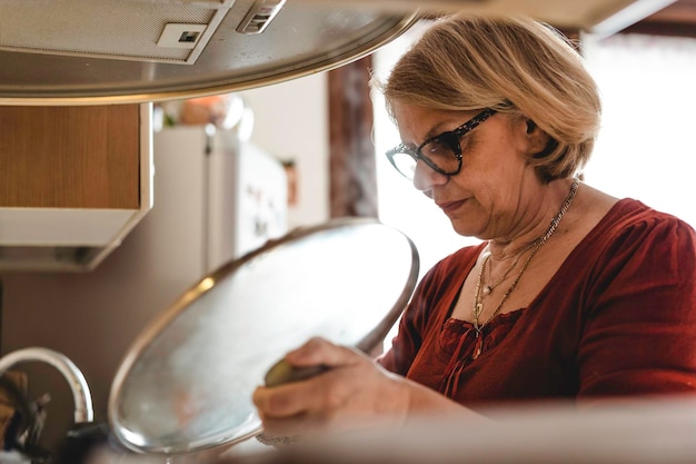 Ältere Frau, die das Mittagessen in der Küche kocht Ältere Frau in der Küche, die den Deckel des Topfes anhebt, um das Kochen zu überprüfen Boomer-Frau, die damit beschäftigt ist, das Mittagessen für die Familie zu kochen