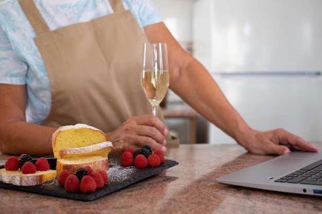 Ältere Frau, die ein Glas Wein hält, während sie auf dem Laptop surft Hausgemachter Pflaumenkuchen, dekoriert mit Beeren auf dem Tisch