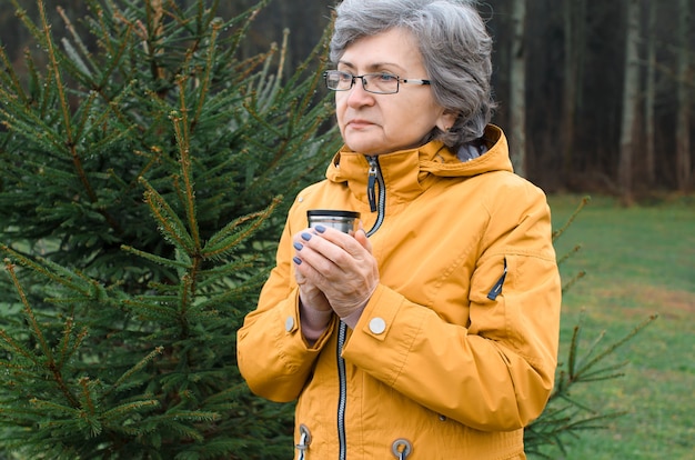 Ältere Frau, die eine Tasse heißen Tee draußen im Wald hält. Ältere Frau, die versucht, in kaltem Wetter warm zu halten.