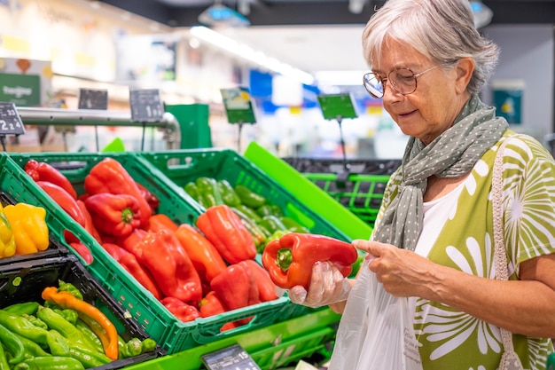Ältere Frau, die frisches Gemüse auswählt, das eine rote Paprika in der Lebensmittelabteilung im Supermarkt-Konsumkonzept hält, steigende Preise Inflation