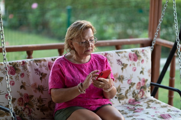 Ältere Frau, die Handy benutzt, während sie auf der Außenschaukel auf der Terrasse sitzt