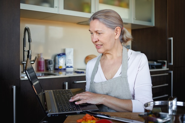 Ältere Frau, die mit Hilfe des Rezepts auf Laptop kocht.