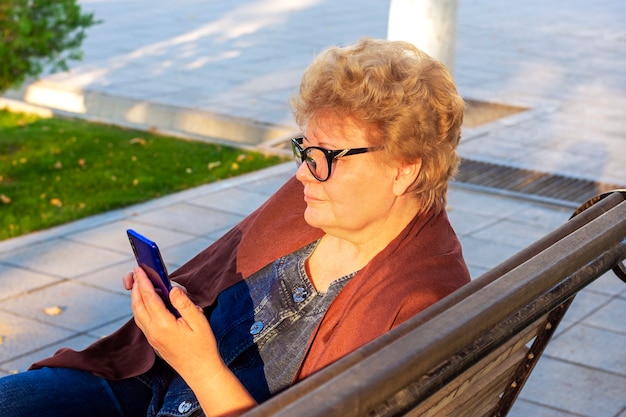 Ältere Frau, die Smartphone im Park auf Bank im Herbstwetter verwendet