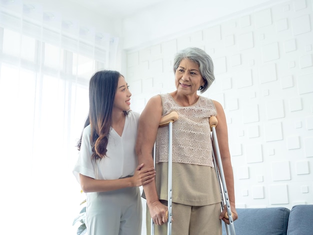 Ältere Frau, die versucht, auf Krücken zu gehen, die von jungen asiatischen Frauen sorgfältig im Aufwachraum gehalten und in den Armen gehalten werden, um alten Frauen zu helfen, Senior-Therapiepatienten im Gesundheitswesen zu Hause zu behandeln
