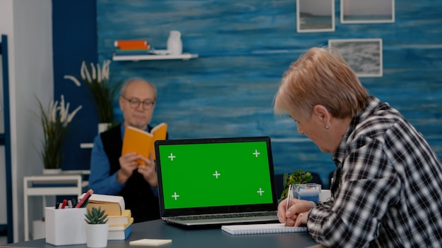 Ältere Frau, die von zu Hause aus auf dem Greenscreen-Monitor arbeitet und Notizen über das Notebook macht