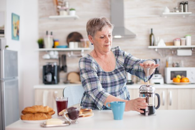 Ältere Frau, die während des Frühstücks Kaffee mit französischer Presse in der Küche kocht. Ältere Menschen genießen morgens frisches braunes Kaffee-Espresso-Koffein aus Vintage-Becher, Filter entspannen Erfrischung