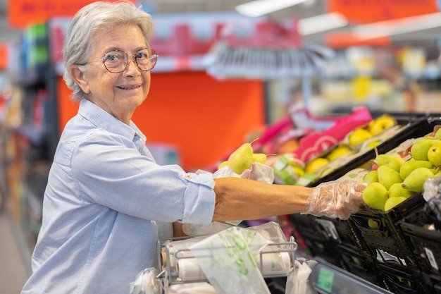 Ältere Frau im Supermarkt, die Handschuhe trägt, um einige Birnen in der Preiserhöhung und Inflation der Obstabteilung auszuwählen