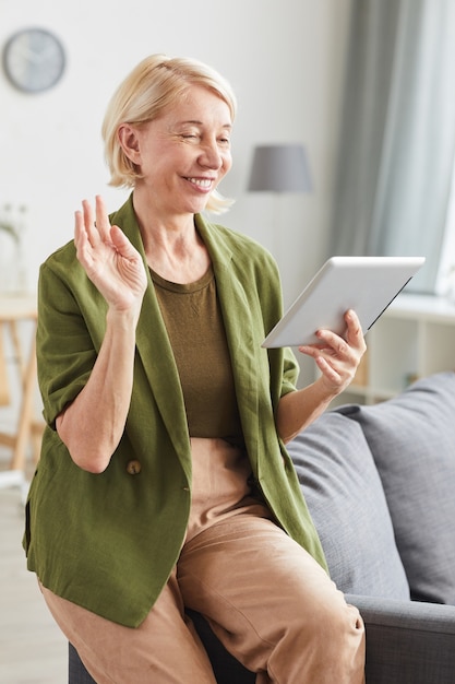 Ältere Frau in der Freizeitkleidung, die digitales Tablett betrachtet und zu ihrem Freund lächelt, während sie zu Hause sitzt