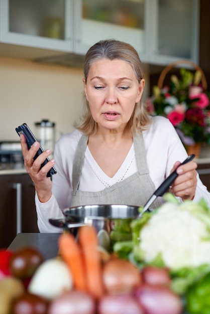 Ältere Frau in der Küche, die Essen vorbereitet und Smartphone hält