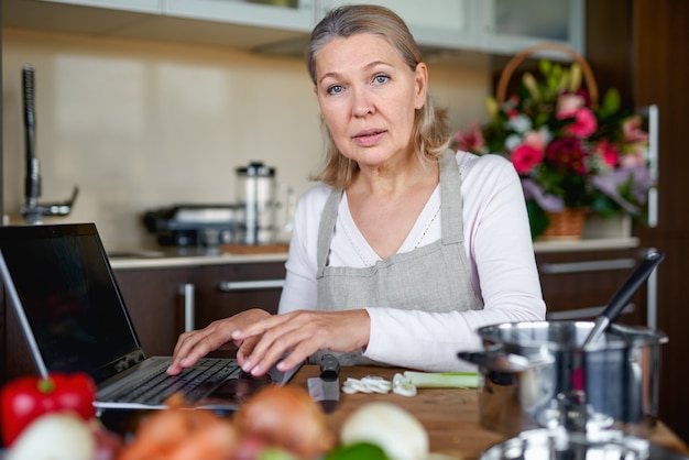 Ältere Frau in der Schürze in der Küche bereitet Essen zu und benutzt Laptop