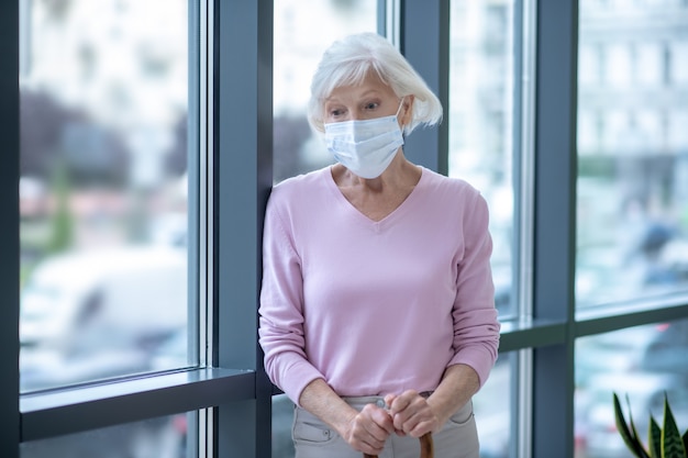 Ältere Frau in einer Gesichtsmaske, die nahe dem Fenster steht