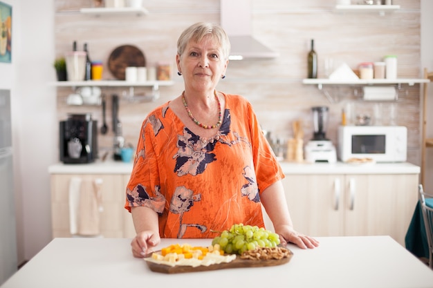 Ältere Frau mit Blick auf die Kamera in der heimischen Küche mit einer Auswahl an leckerem Käse und Trauben auf der Tischplatte.