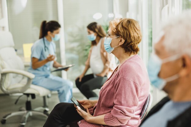 Ältere Frau mit Gesichtsmaske, die vor der Covid-19-Impfung in einem Wartezimmer eines Krankenhauses sitzt.