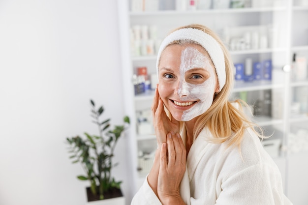 Ältere Frau mit kosmetischer Maske auf halbem Gesicht im Schönheitssalon.