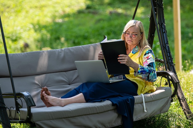 Ältere Frau mit Laptop und Dokumenten, die im Garten auf einem grünen Home-Office-Konzept der Schaukelcouch arbeiten