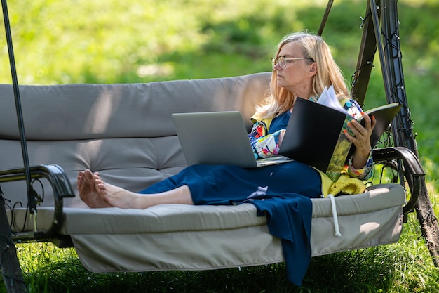 Ältere Frau mit Laptop und Dokumenten, die im Garten auf einem grünen Home-Office-Konzept der Schaukelcouch arbeiten