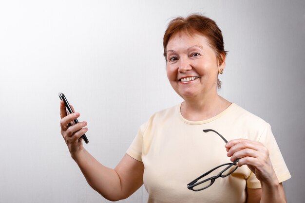 Ältere Frau schaut auf das Telefon mit Brille auf Weiß in einem hellen T-Shirt