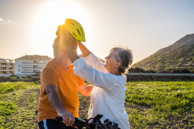 Ältere Frau setzt ihrem Mann einen Schutzhelm auf, um Fahrrad zu fahren. Helles Sonnenuntergangslicht