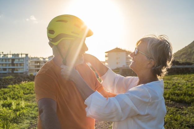 Ältere Frau setzt ihrem Mann einen Schutzhelm auf, um Fahrrad zu fahren. Helles Sonnenuntergangslicht