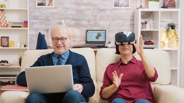 Ältere Frau sitzt auf dem Sofa und trägt eine Virtual-Reality-Brille. Alter Mann sitzt auf dem Sofa mit Laptop neben seiner Frau