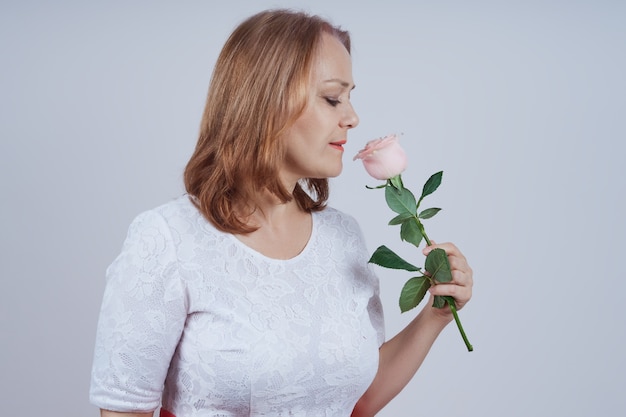 Ältere Frau steht im Profil, in einem weißen Kleid, das eine Rosenblume hält. in über graue Wand.