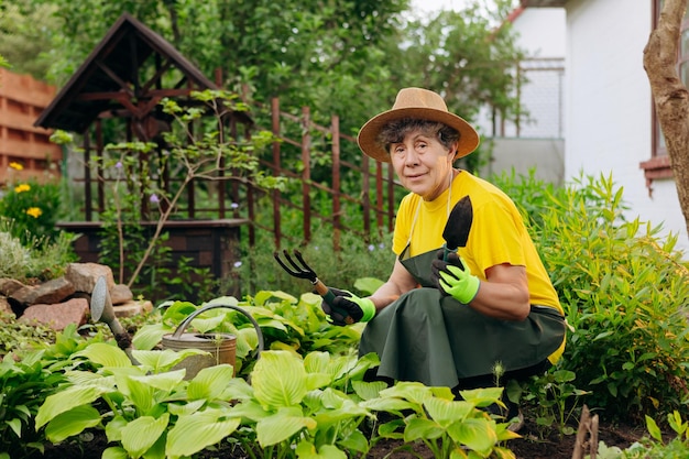 Ältere Gärtnerin mit Hut, die in ihrem Garten mit Arbeitswerkzeugen arbeitet Das Konzept der Gartenarbeit, das Wachsen und Pflegen von Blumen und Pflanzen