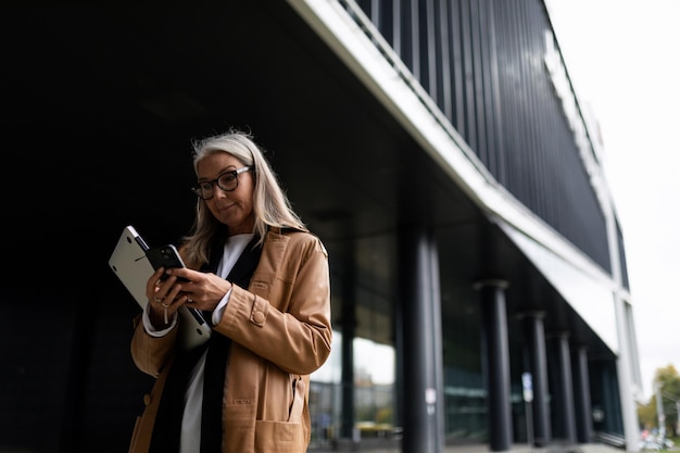 Ältere Geschäftsfrau mittleren Alters schaut auf das Telefon, das er mit einem Laptop in der Hand neben dem hat