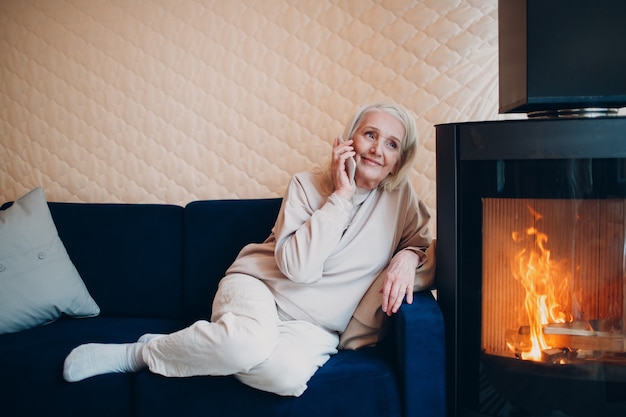 Ältere grauhaarige Frau, die auf Sofa sitzt und Smartphone im Wohnzimmer mit Kaminkamin spricht