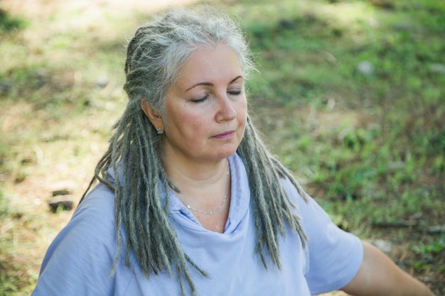 Ältere grauhaarige Frau mit Dreadlocks im Sitzen mit geschlossenen Augen im Freien, Meditation und Daydr