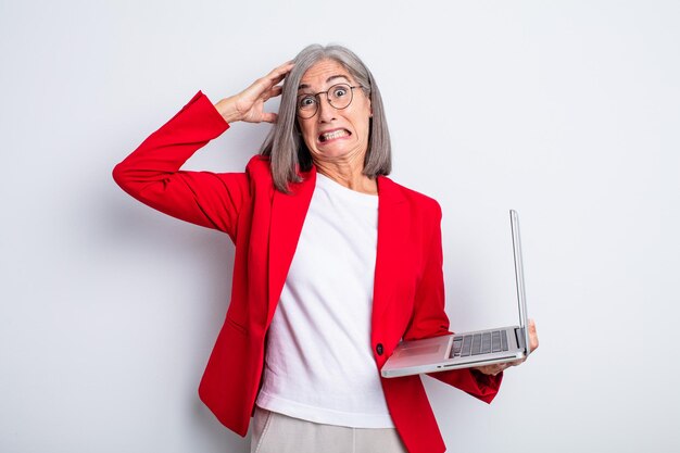 Ältere hübsche Frau, die sich gestresst, ängstlich oder verängstigt fühlt, mit den Händen auf dem Kopf. Business- und Laptop-Konzept