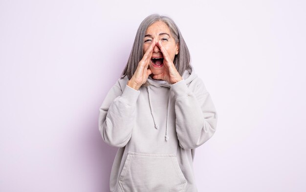 Ältere hübsche Frau, die sich glücklich, aufgeregt und positiv fühlt, mit den Händen neben dem Mund einen großen Schrei ausstößt und ruft